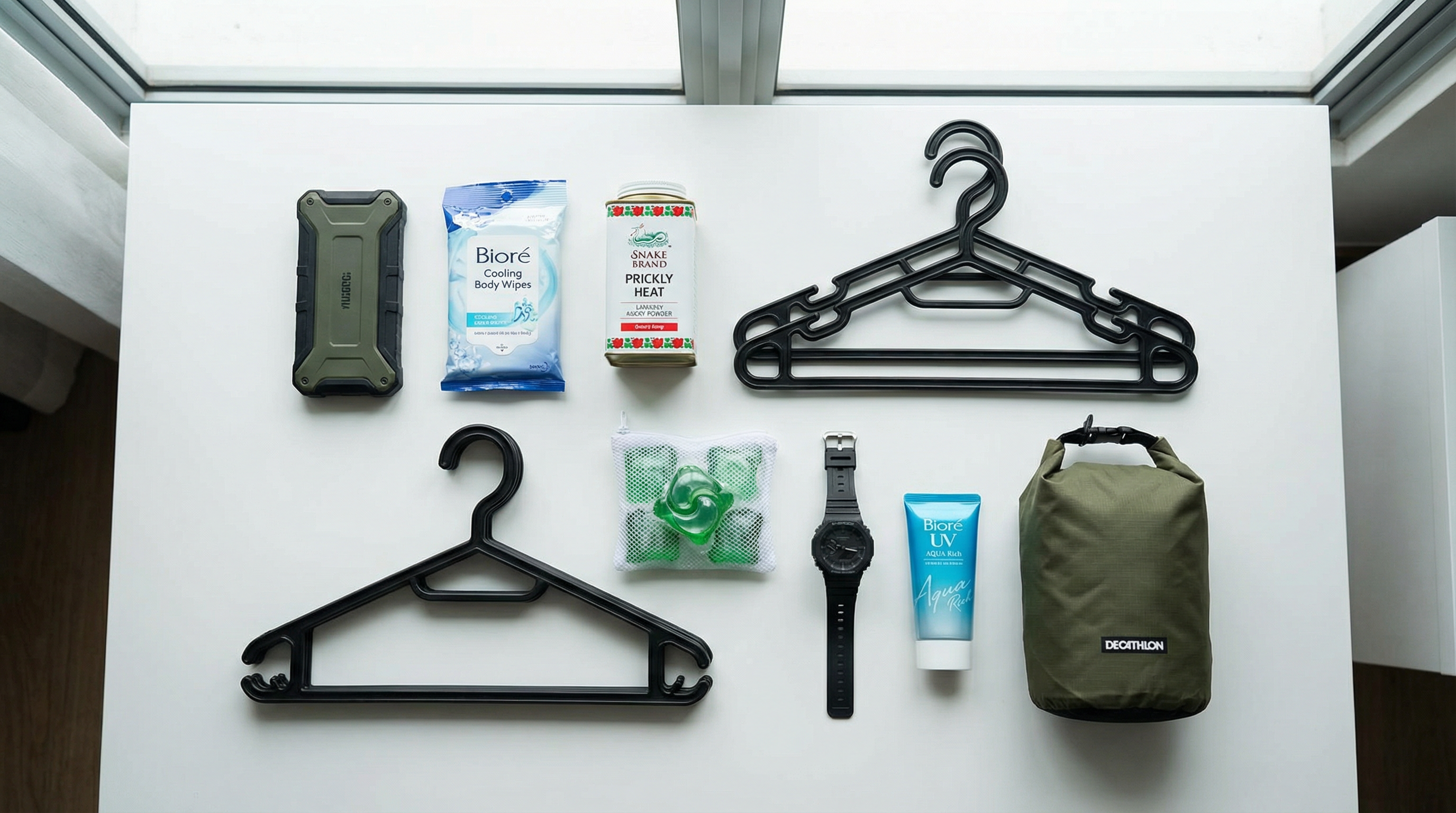 Flat lay of essential BMT packing items including power bank, wet wipes, prickly heat powder, hangers, and watch